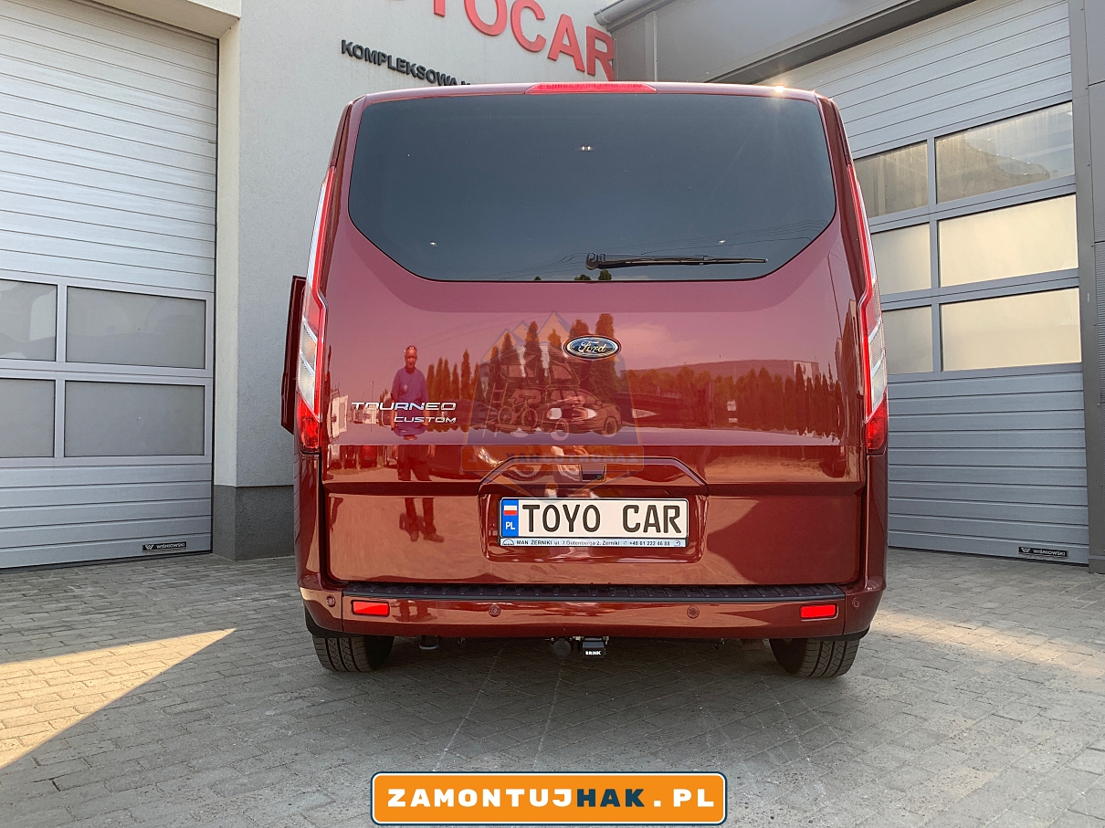 Zdjęcie przedstawia Forda Tourneo Custom z zamontowanym hakiem holowniczym wypinanym pionowo brink, zdjęcie zostało zrobione od tyłu na tle serwisu zamontujhak.pl